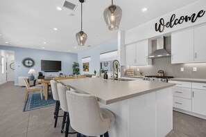 A modern kitchen with a large island, bar stools, a dining area with a TV, and a 'Welcome' sign. - El Encanto: EV Charger, Pool, Spa & BBQ (La Quinta)