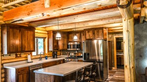 Eine rustikale Kücheneinrichtung in einem Blockhaus mit Holzausbau, einer zentralen Kochinsel mit Barhocker, Edelstahlgeräten und hängenden Pendelleuchten.