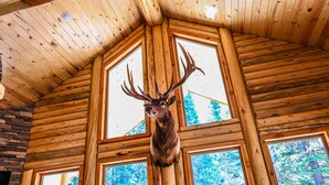 Ein Holzblockhaus-Innenraum mit einem aufgehängten Hirschkopf, großen Fenstern und einem Kamin aus Stein.