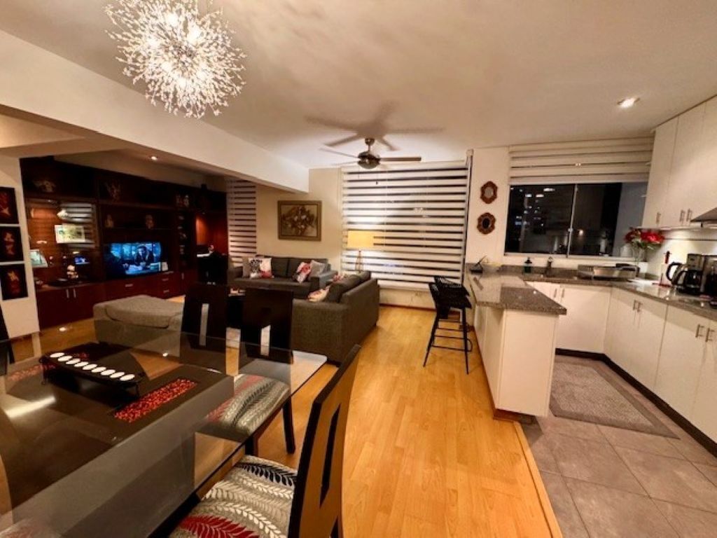 En moderne stue med sofa, spisebord og tv. Der er et køkkenområde med skabe og et vindue.