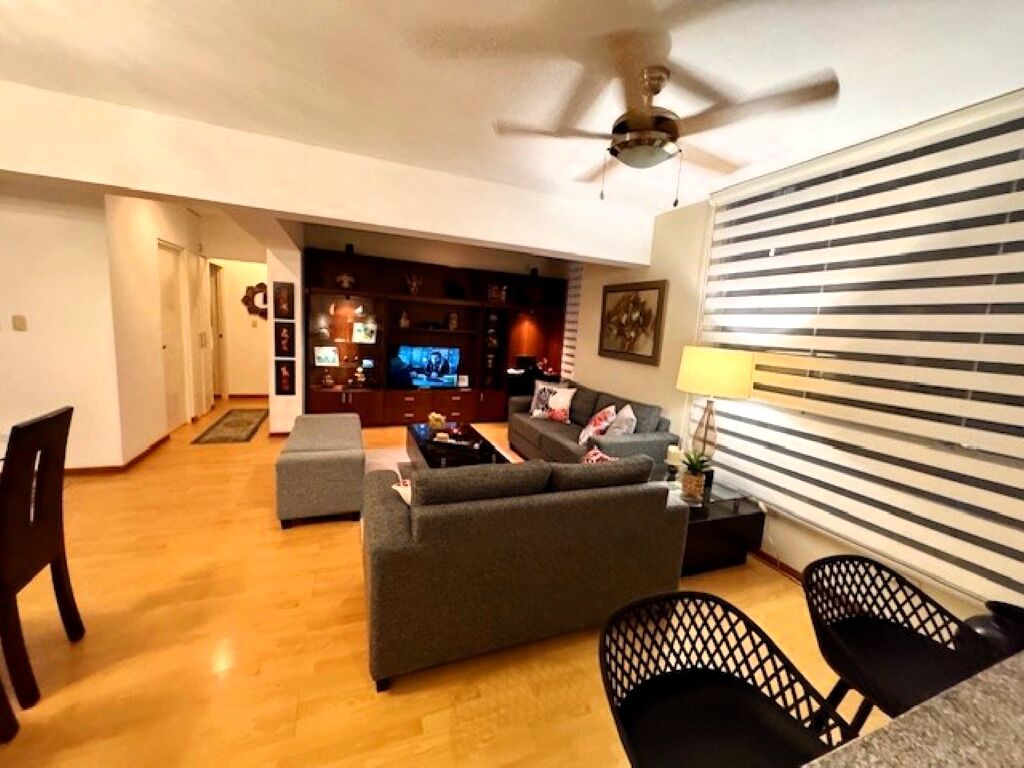 Un salon avec un canapé, une table basse et un téléviseur. On y trouve également un ventilateur de plafond, une lampe et une fenêtre avec des stores.