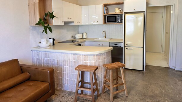 Una cocina moderna con una isla central, taburetes de madera, un microondas, un refrigerador y una lámpara colgante.