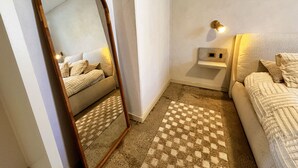 Un dormitorio con un espejo grande, una mesita de noche con lámpara, una alfombra a cuadros y una cama con almohadas.