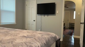 Une chambre à coucher avec un lit, un téléviseur fixé au mur, une porte et un couloir menant à une autre pièce.