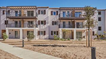 A modern apartment building with multiple balconies, a sandy ground, and a clear blue sky.
