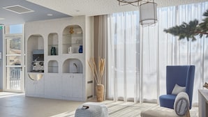 Ein modernes Wohnzimmer mit blauem Sessel, weißem Pouf und einer Küchenzeile mit weißen Schränken und einer integrierten Regalanlage.