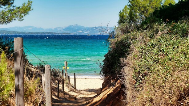 Un sentier sablonneux qui longe la plage et mène à une étendue de sable bordée d’une mer d’un bleu limpide et dominée par des montagnes lointaines.