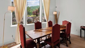 A dining room with a large wooden table, six red upholstered chairs, a floor lamp, and a view of a mountainous landscape through a window with beige curtains. - Home in Midway - Short Drive to Deer Valley Gondola | Mountain Views | Large Deck | Private Bedrooms (Midway)
