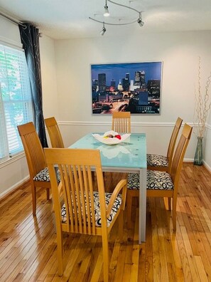 A dining area with a glass-top table, four wooden chairs with patterned cushions, a cityscape painting on the wall, and a vase with branches.