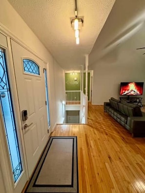 A hallway with a wooden floor, a door with a glass panel, a staircase, and a television on the right.