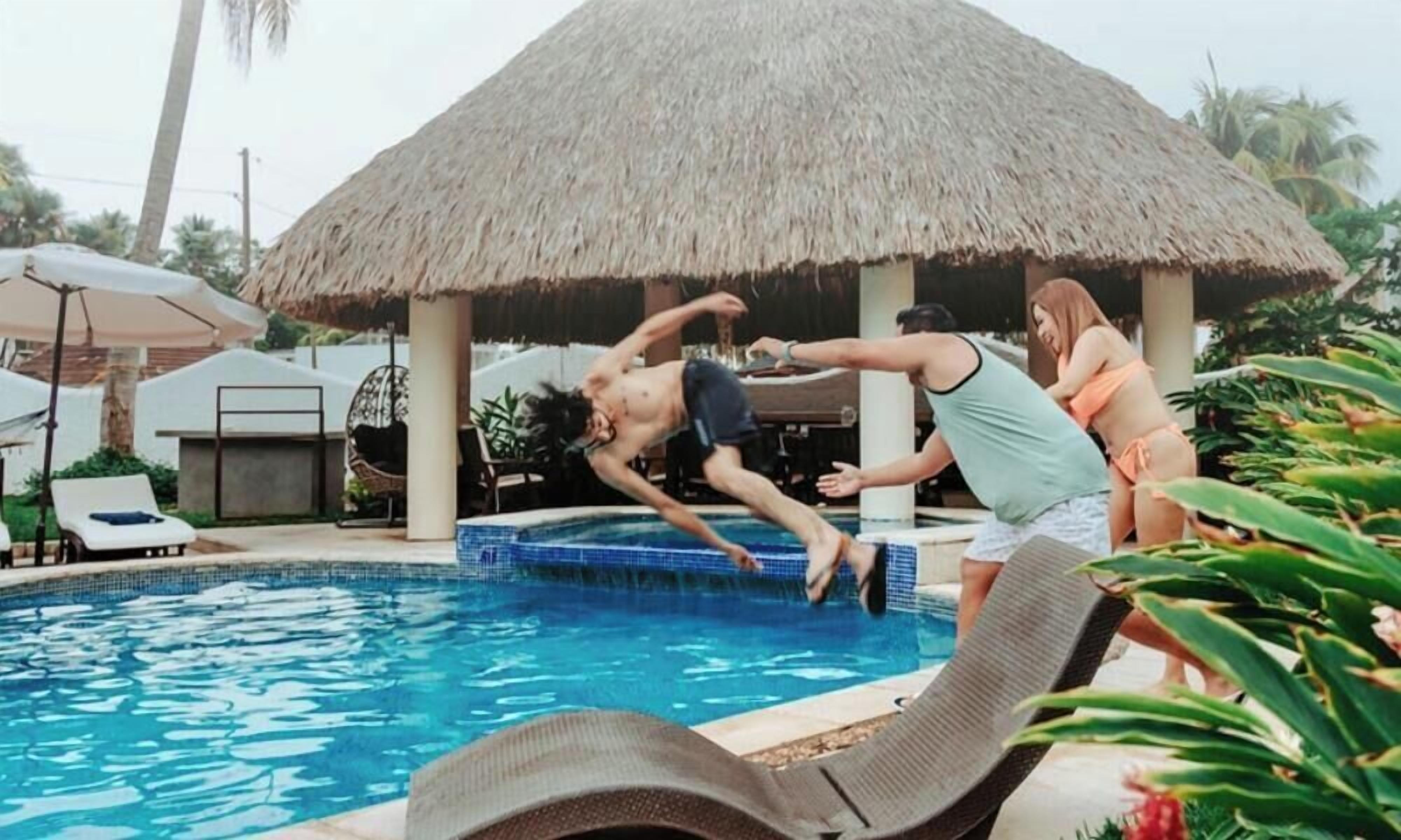 A swimming pool with a thatched roof structure, lounge chairs, and palm trees in the background.