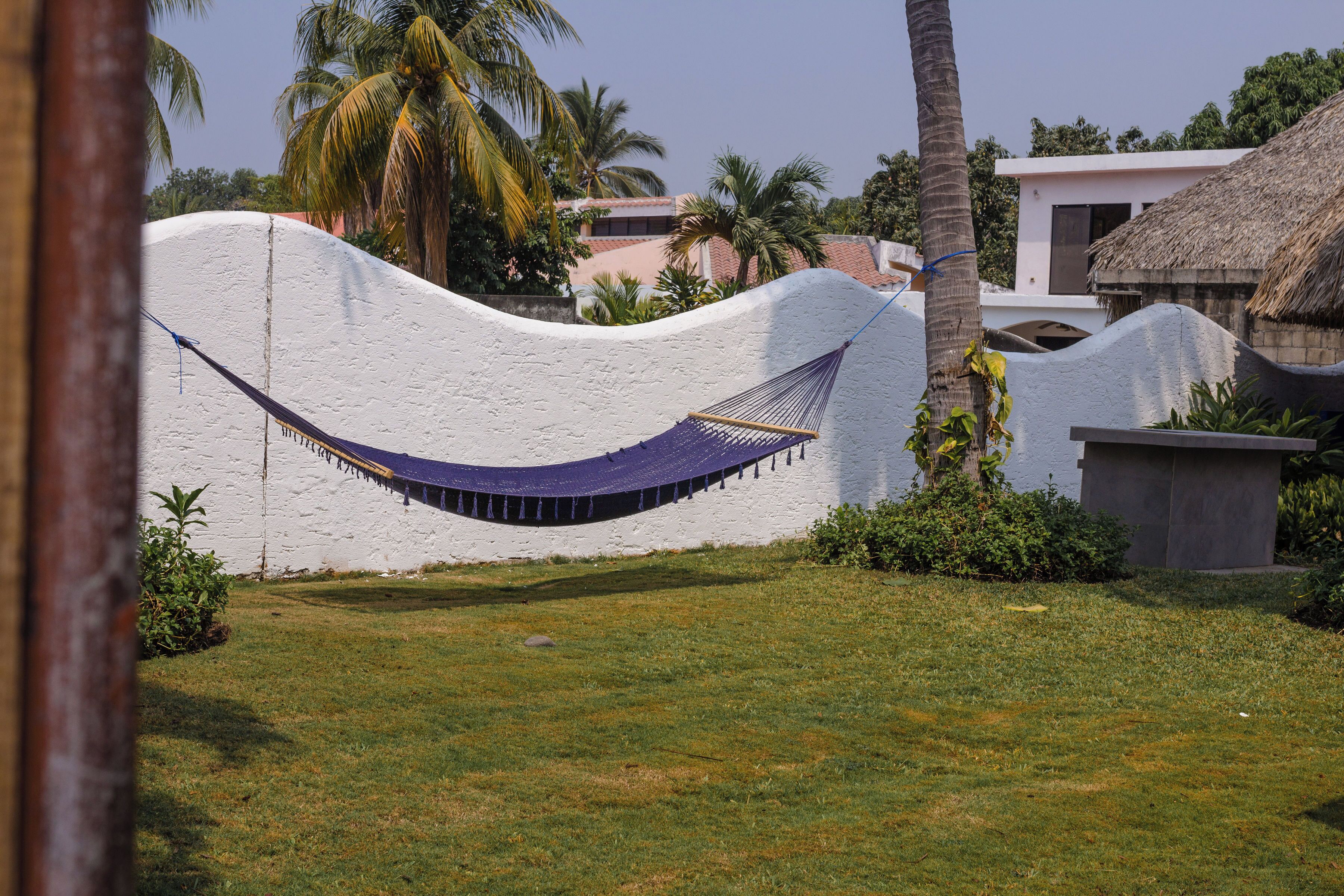 A hammock tied between a white wall and a palm tree, with a grassy area and a building with a thatched roof in the background.