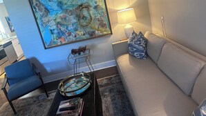 Una sala de estar con un sillón azul, un sofá color beige, una mesita de centro con objetos decorativos, un cuadro en la pared y una lámpara.