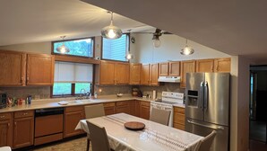 A modern kitchen with wooden cabinets, a stainless steel refrigerator, a dining table with chairs, and pendant lighting.
