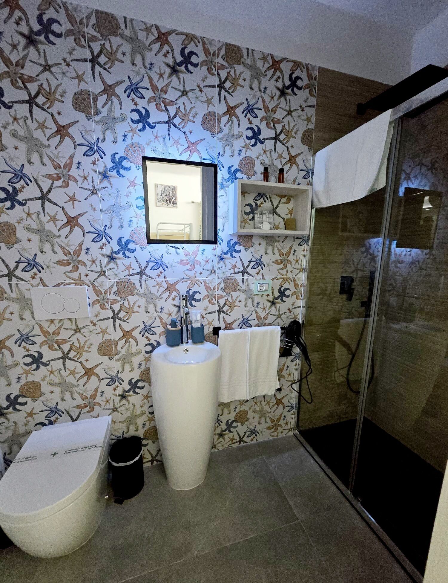 A bathroom with a sea-themed wallpaper, a sink, a toilet, and a glass shower enclosure.