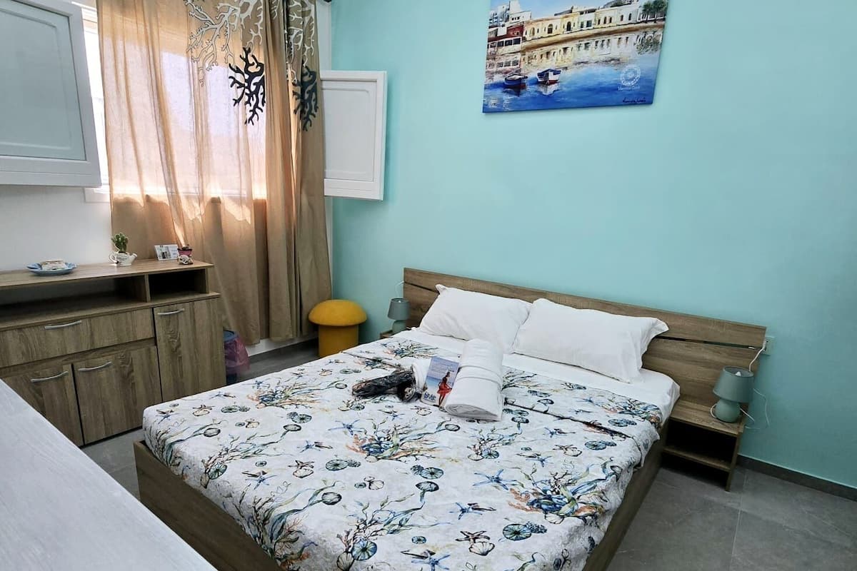 A bedroom with a bed, a bedside table, a nightstand, a window with curtains, a painting on the wall, and a small aquarium on the floor.