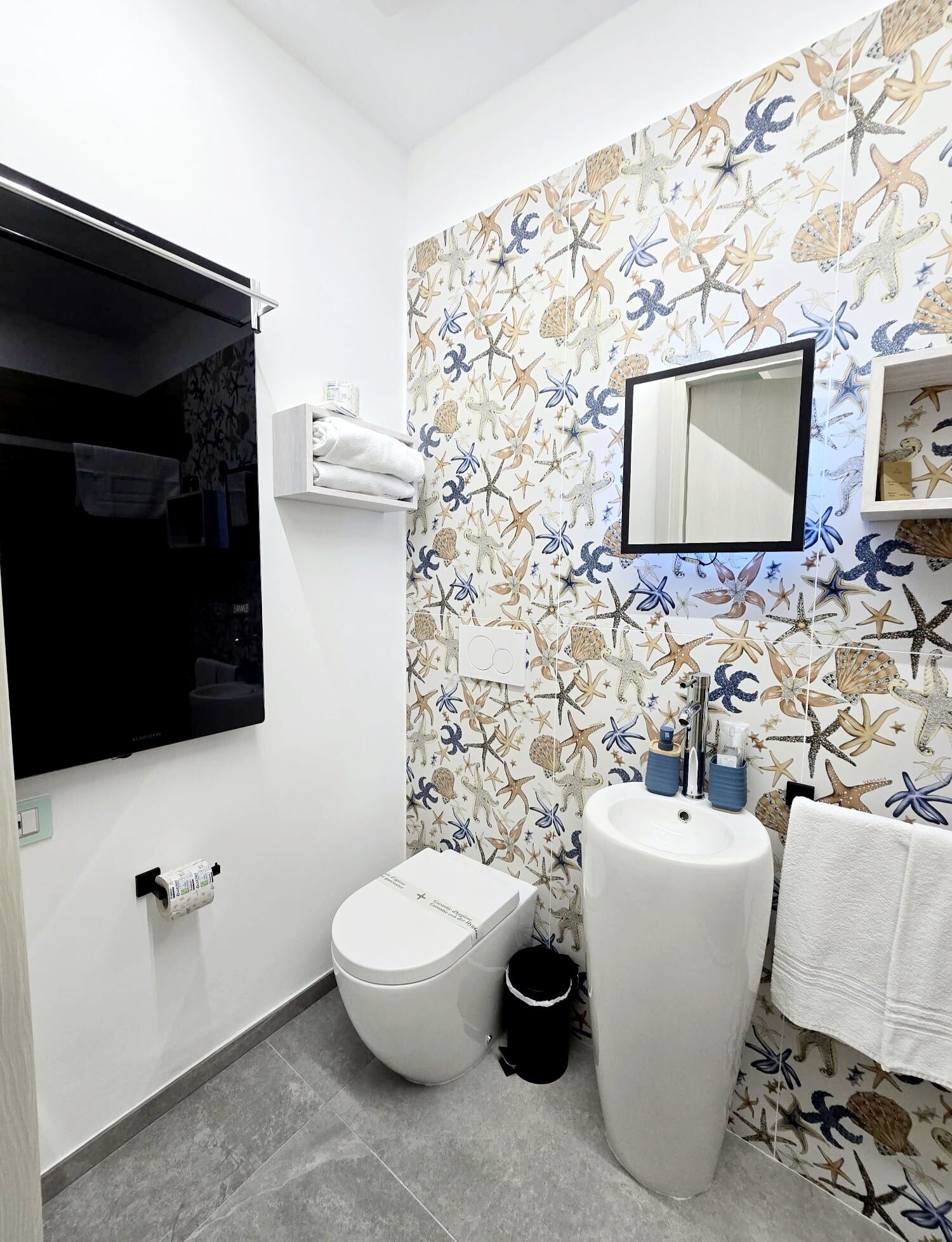 Ein Badezimmer mit Toilette, Waschbecken und Spiegel. Die Wände sind mit einer Meeres-Tapete dekoriert.