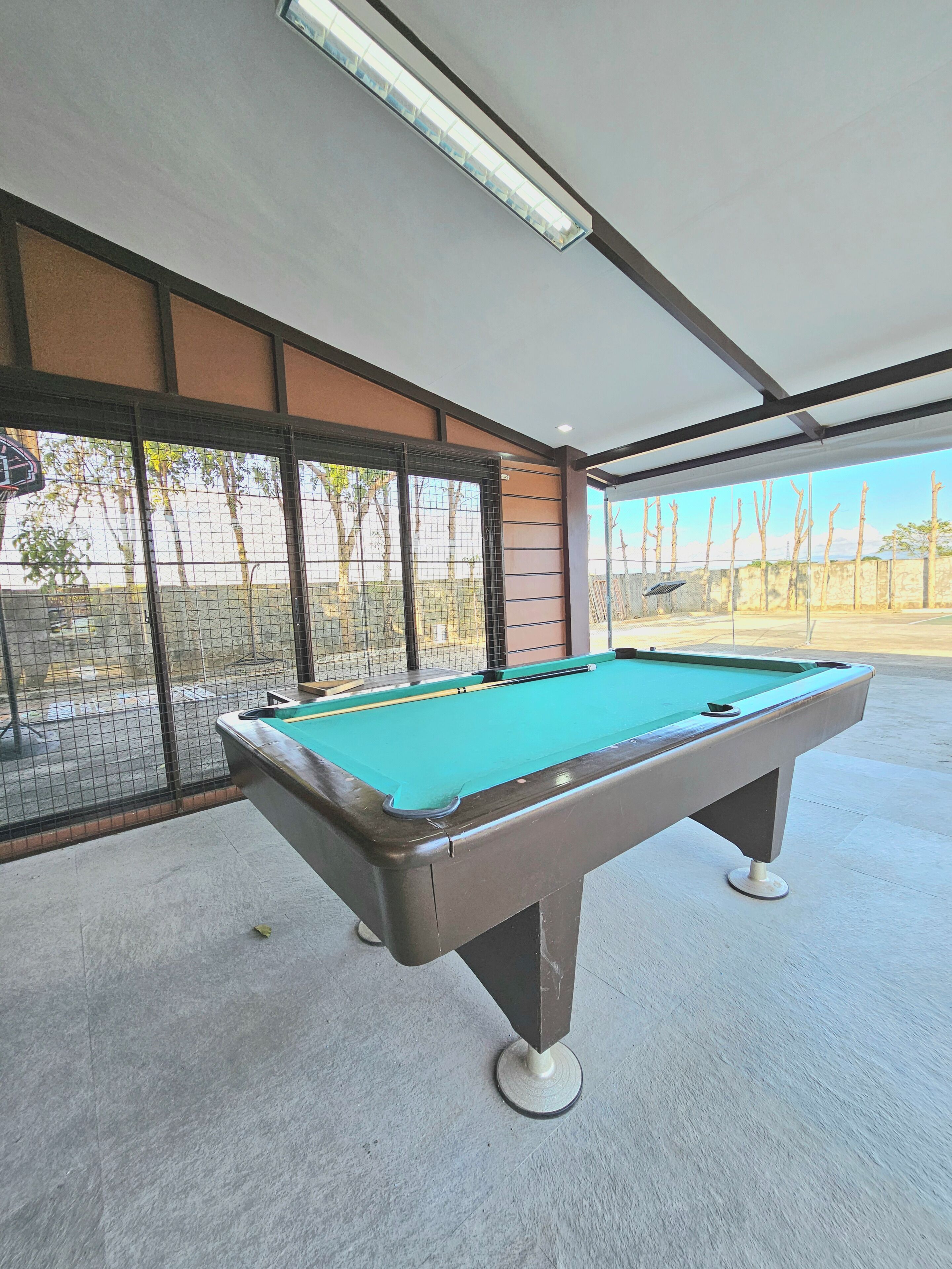 Una mesa de billar en una sala con ventanas amplias, una lámpara de techo y vistas a árboles y una cerca afuera.