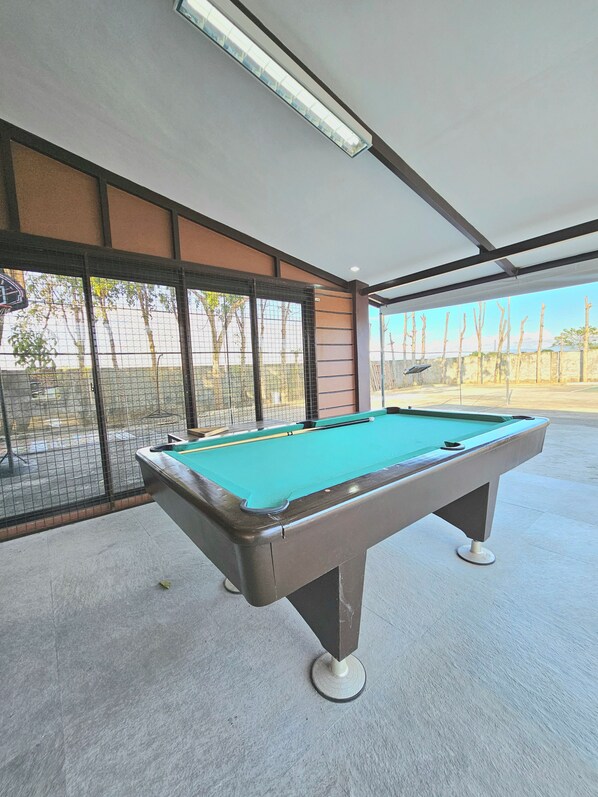 A pool table in a room with large windows, a ceiling light, and a view of trees and a fence outside.