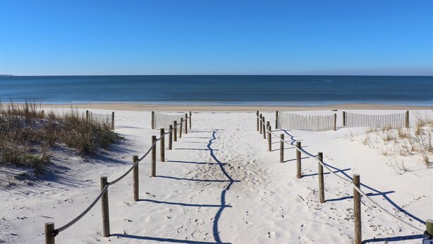 Ein sandiger Küstenweg führt zum Meer, abgegrenzt durch Holzbeschilderung und Seile.