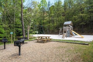 A playground with swings and a slide, surrounded by trees.