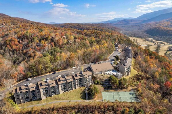 A resort with multiple buildings, a tennis court, and a parking area surrounded by autumn foliage.