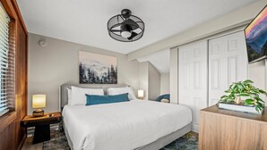 Habitación de hotel con cama, mesita de noche, lámpara, cuadro, planta y puerta.