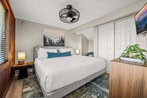 A hotel room with a bed, a nightstand, a lamp, a painting, a plant, and a door.