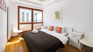 Een moderne slaapkamer met een groot bed, witte nachtkastjes, een witte stoel en een raam met uitzicht op gebouwen.