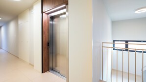 Un ascenseur moderne avec un revêtement mural en bois, un couloir épuré aux carreaux de couleur claire et une rampe métallique sur un escalier.