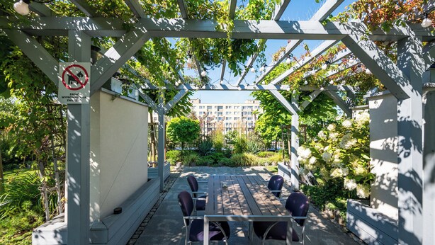 En utomhusplats med sittmöbler, ett pergola-tak, bord och stolar, omgiven av grönska och med utsikt över en byggnad i bakgrunden.