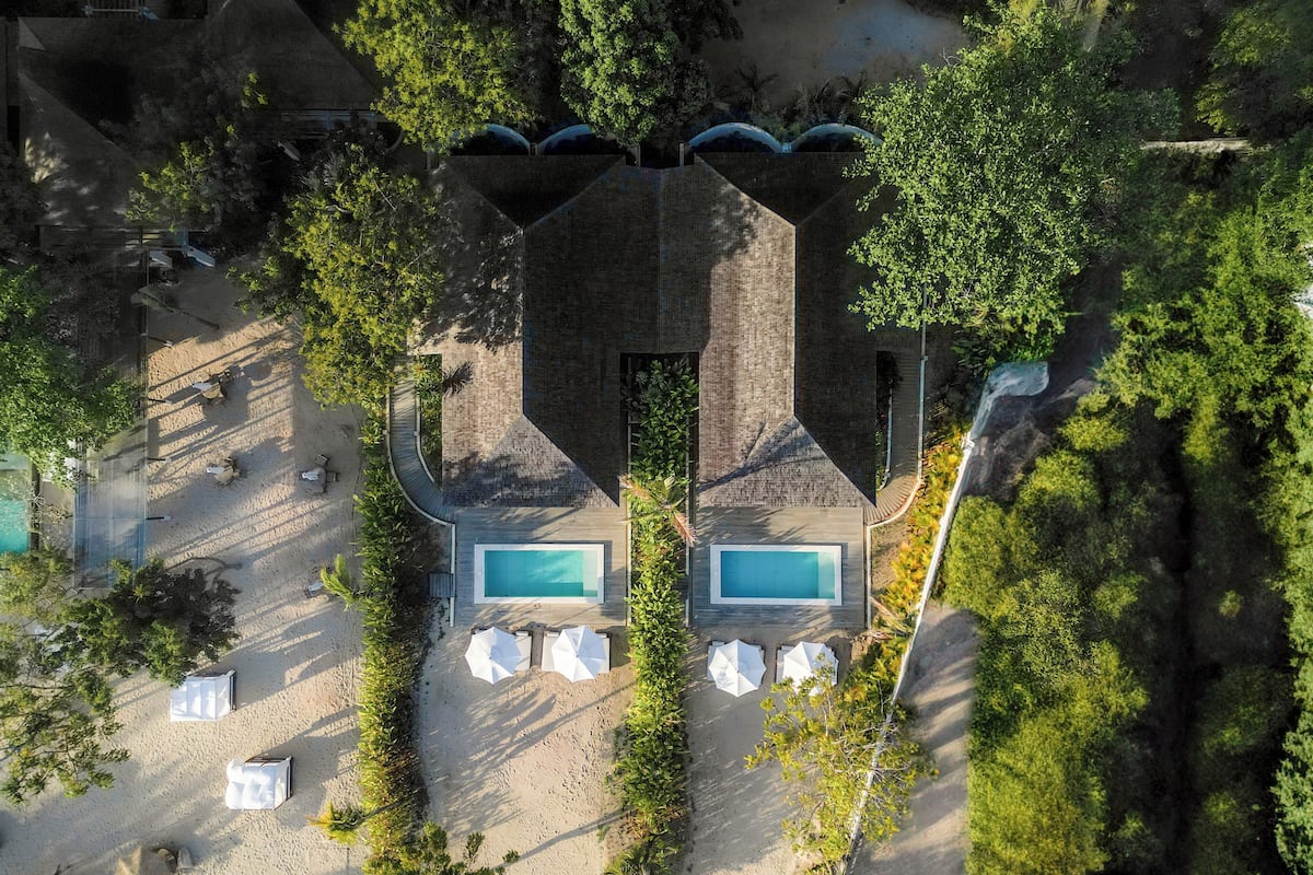 An aerial view of a resort with two swimming pools, surrounded by lush greenery and a sandy area with umbrellas.
