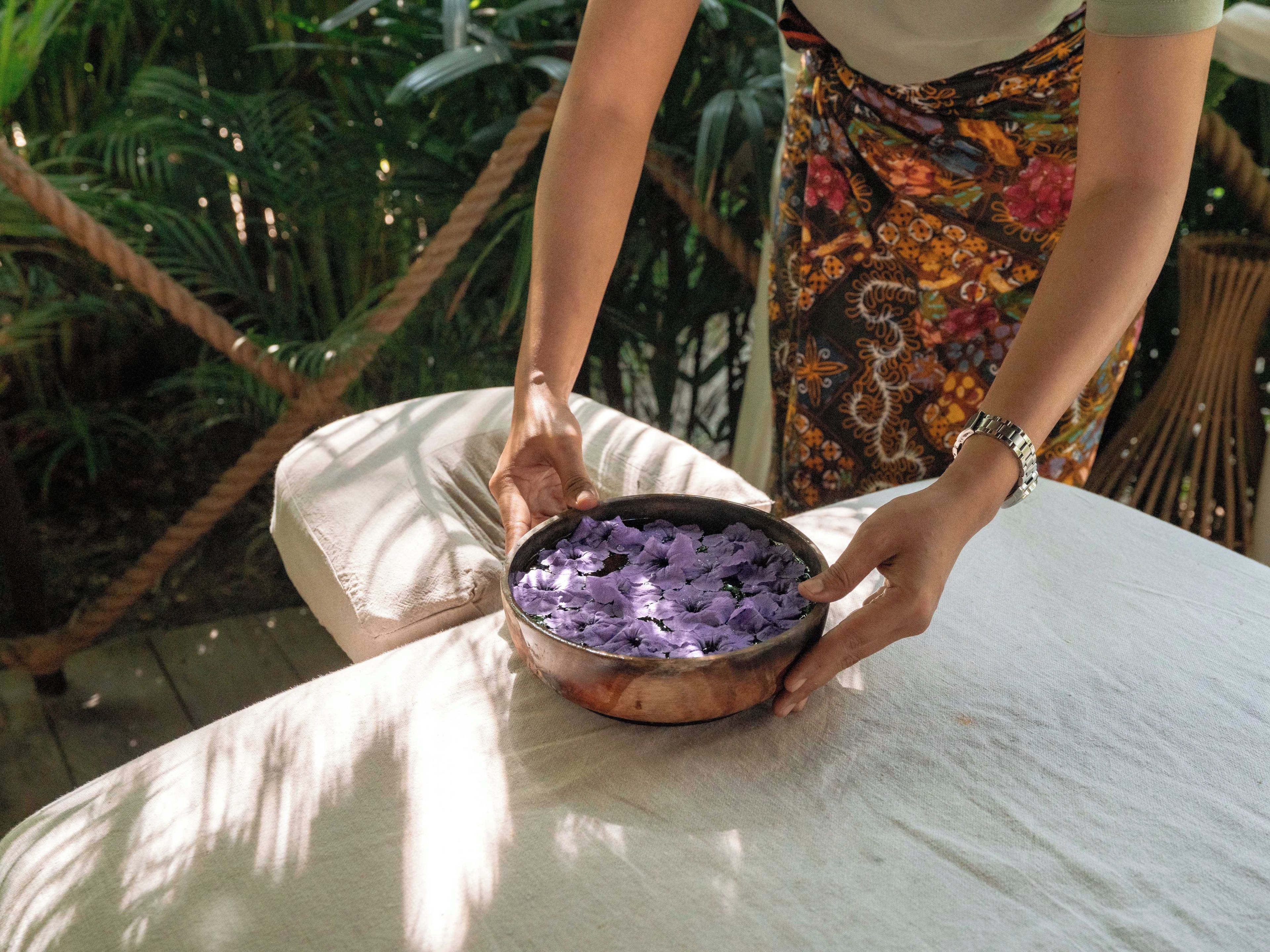Eine Person hält eine Schale mit violetten Blumen, sitzt draußen an einem Tisch mit Grünpflanzen im Hintergrund.