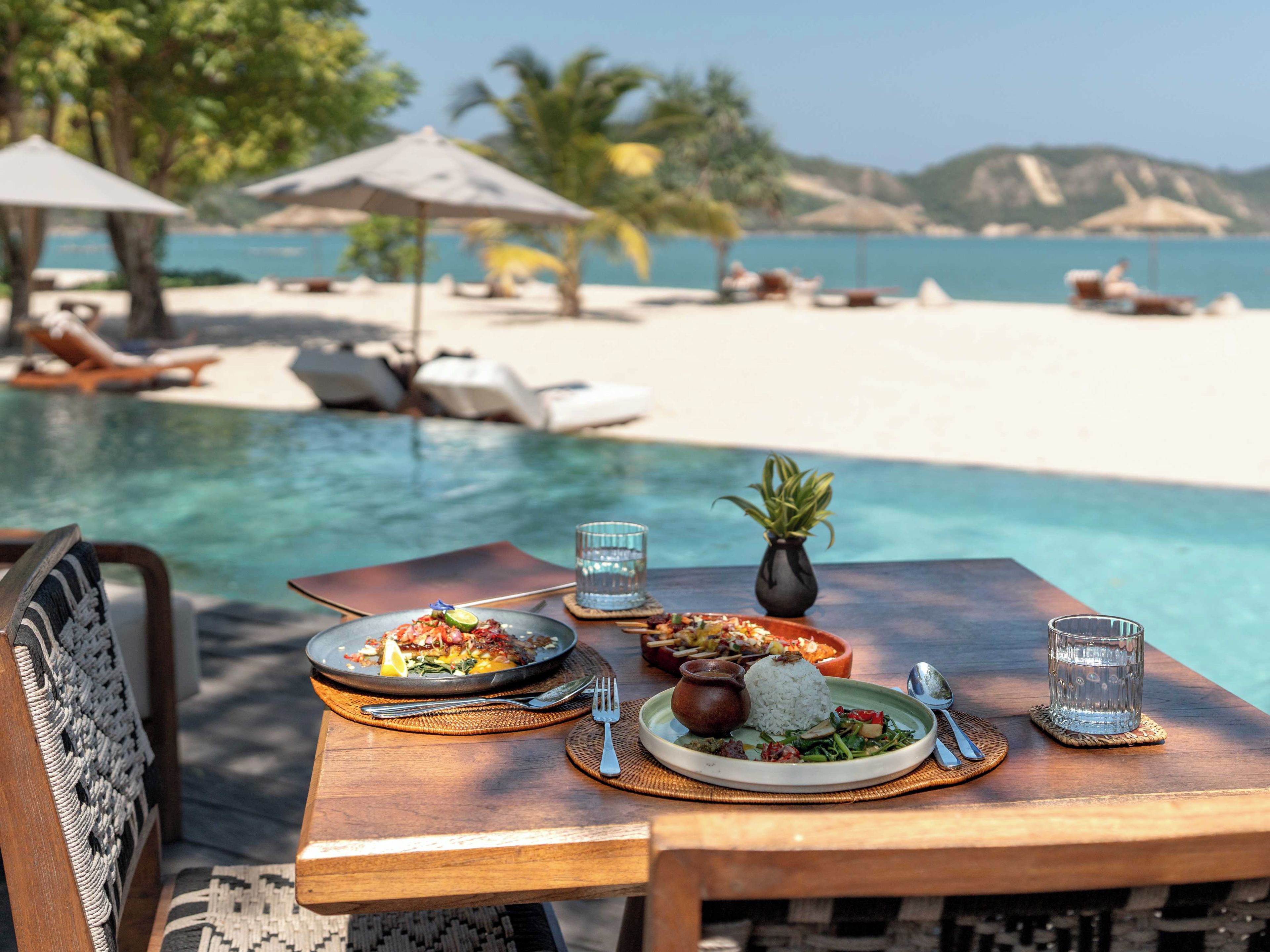 Una mesa de comedor con platos de comida, un vaso de agua y vista a una playa y piscina.