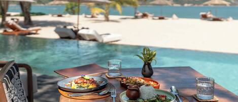 Una mesa de comedor con platos de comida, un vaso de agua y vista a una playa y piscina.