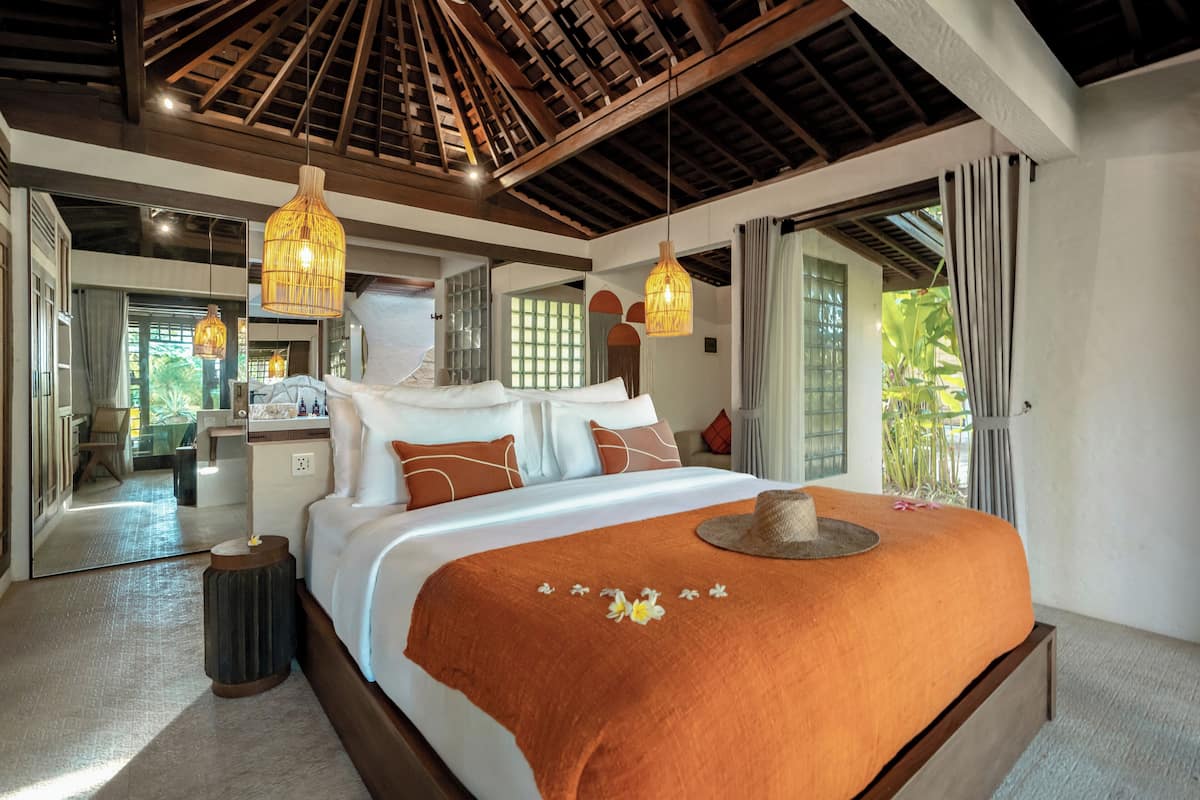 A spacious bedroom with a large bed, multiple hanging lights, and a view of a lush garden through the window.