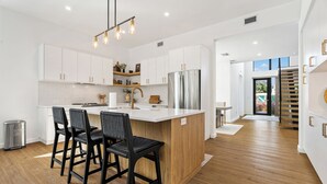 A modern kitchen with white cabinets, a central island with bar stools, a stainless steel refrigerator, and pendant lighting.