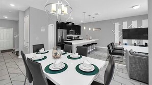 Una cocina moderna con una isla central, zona de comedor con una mesa blanca dispuesta para cuatro personas y un salón con sofá y televisor.
