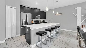 Una cocina moderna con armarios oscuros, una isla blanca con taburetes de bar, electrodomésticos de acero inoxidable e iluminación colgante.