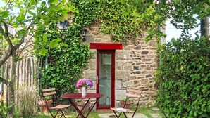 Ein Steinhaus mit einer roten Tür, einem kleinen runden Tisch mit einem Blumenstrauß und zwei Holzsesseln.