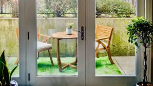 Vista a través de una puerta de vidrio a un patio con una mesa redonda, dos sillas de madera y una planta en maceta.
