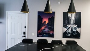 Un salón de conferencias con una mesa de mármol negro, tres lámparas colgantes negras y dos cuadros con vistas urbanas en la pared.