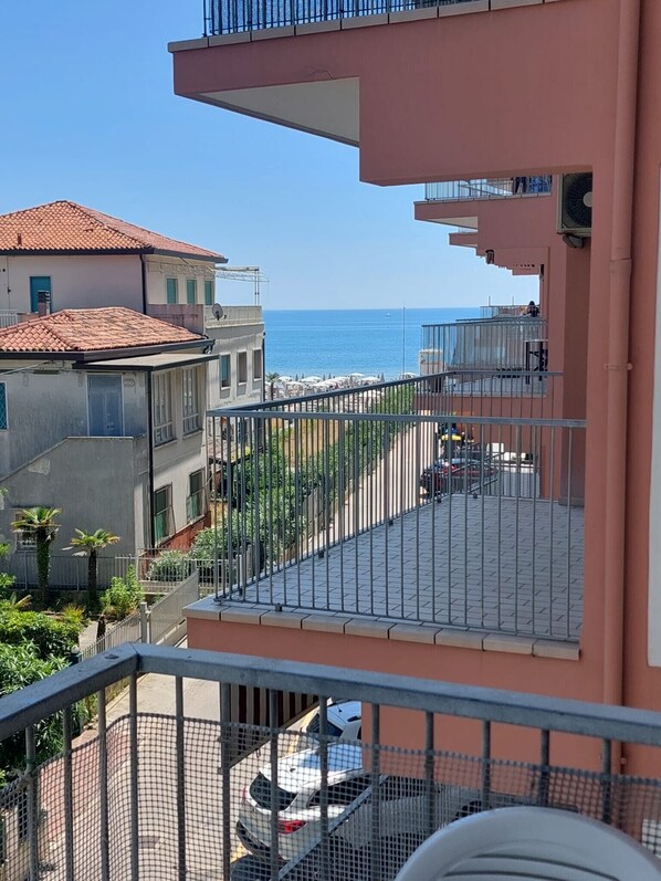 A balcony with a view of the sea, a parking area, and neighboring buildings.