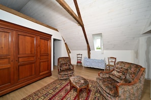 Quarto com teto alto, vigas de madeira, um guarda-roupa espaçoso, duas poltronas estampadas, uma mesinha e uma janela.