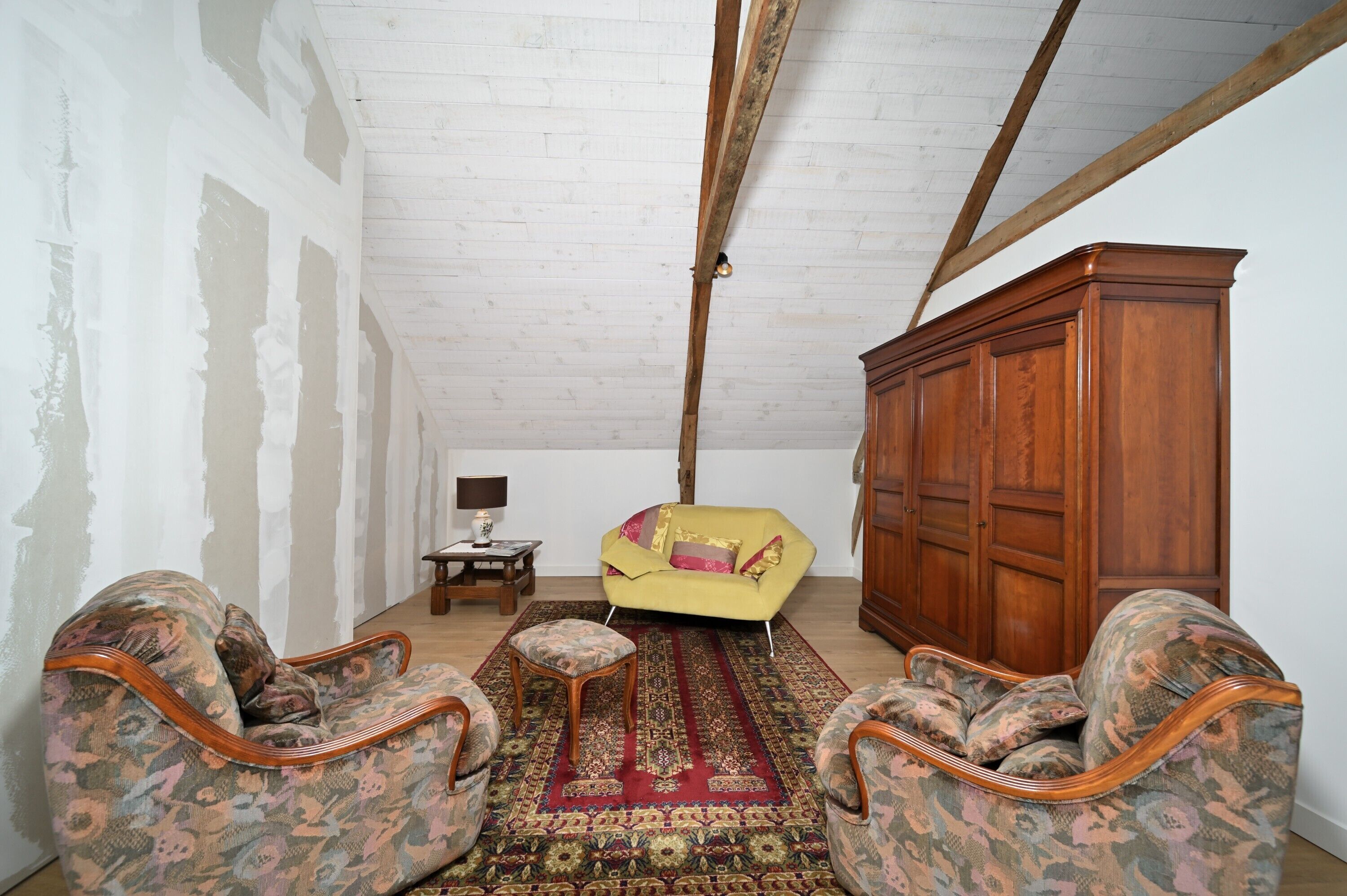 Een kamer met een hoge plafond, houten balken, een kamerbreed tapijt met dessin, een gele bank en twee fauteuils met dessin.