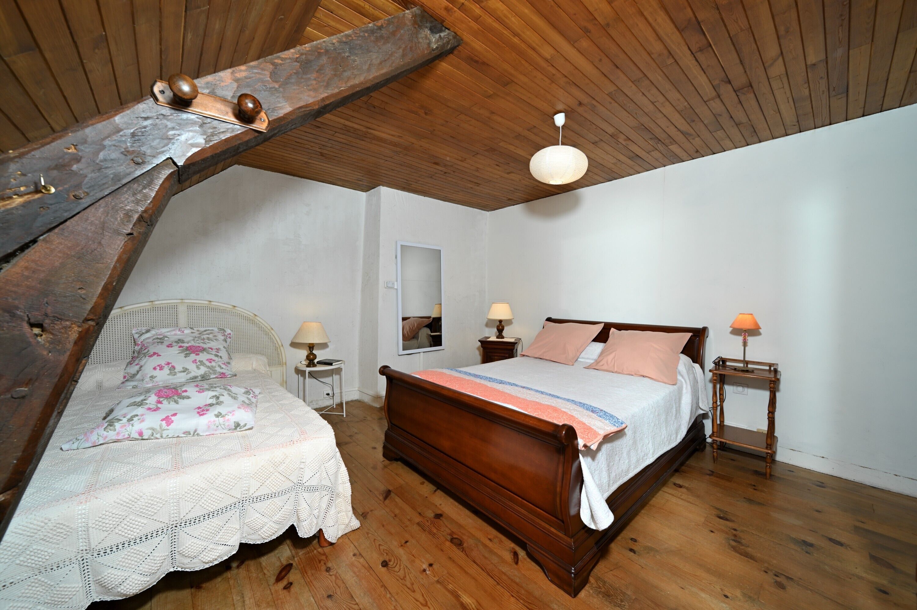 Une chambre avec un plafond en bois, un lit avec un cadre en bois, deux tables de chevet avec des lampes et un mur blanc orné d’un miroir.
