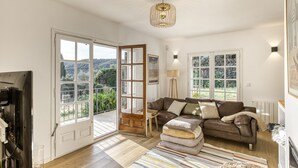 A spacious living room with a brown leather sofa, a wooden coffee table, and a large window offering a view of greenery. - Villa 'Les Aulines' with Mountain View, Wi-Fi and Air Conditioning (Calonge)