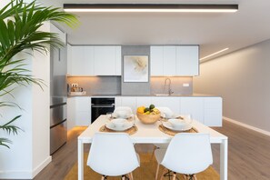 Une cuisine moderne avec des meubles blancs, une table à manger dressée pour deux personnes et un panier de fruits posé sur la table.