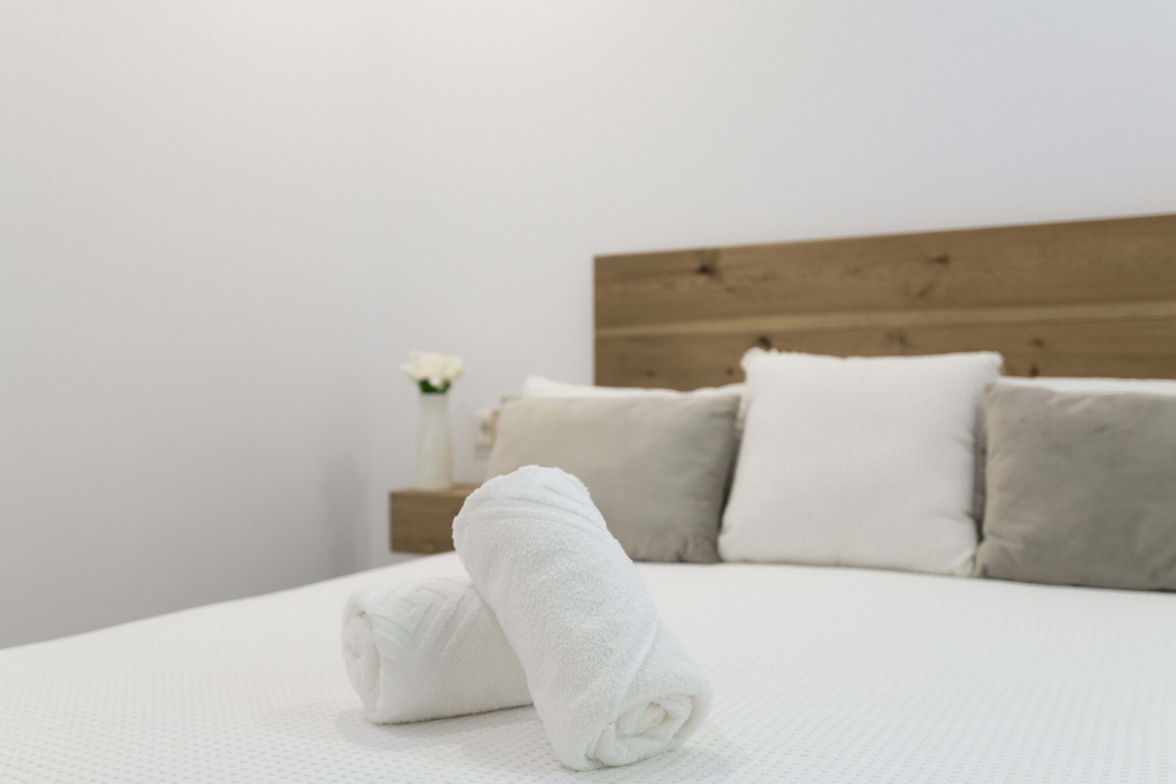 Een opgemaakt bed met witte lakens, een houten hoofdbord en een vaas met witte bloemen op een nachtkastje.