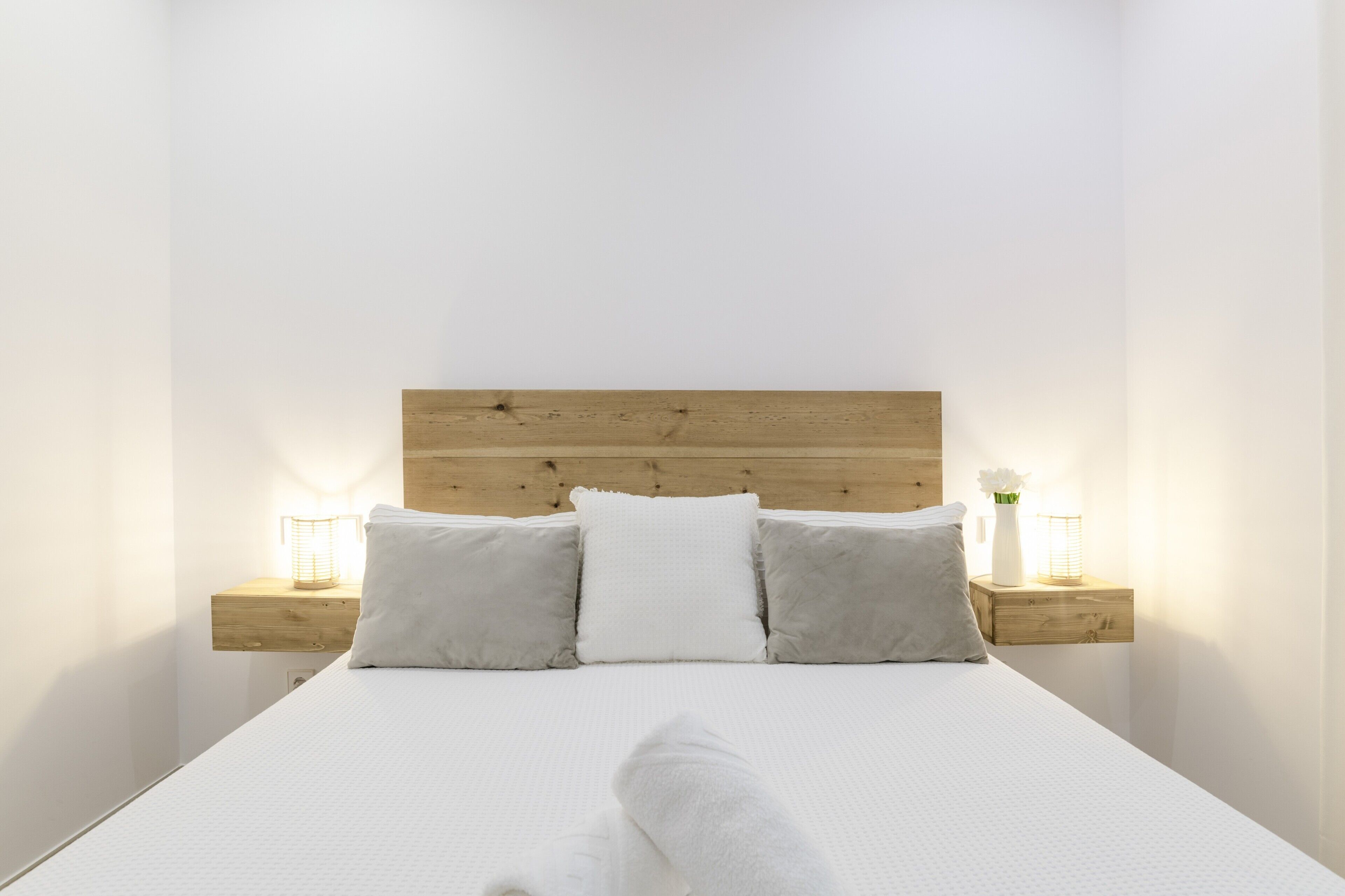 Cama bem arrumada com cabeceira de madeira, duas mesas de cabeceira com abajures e uma parede branca.